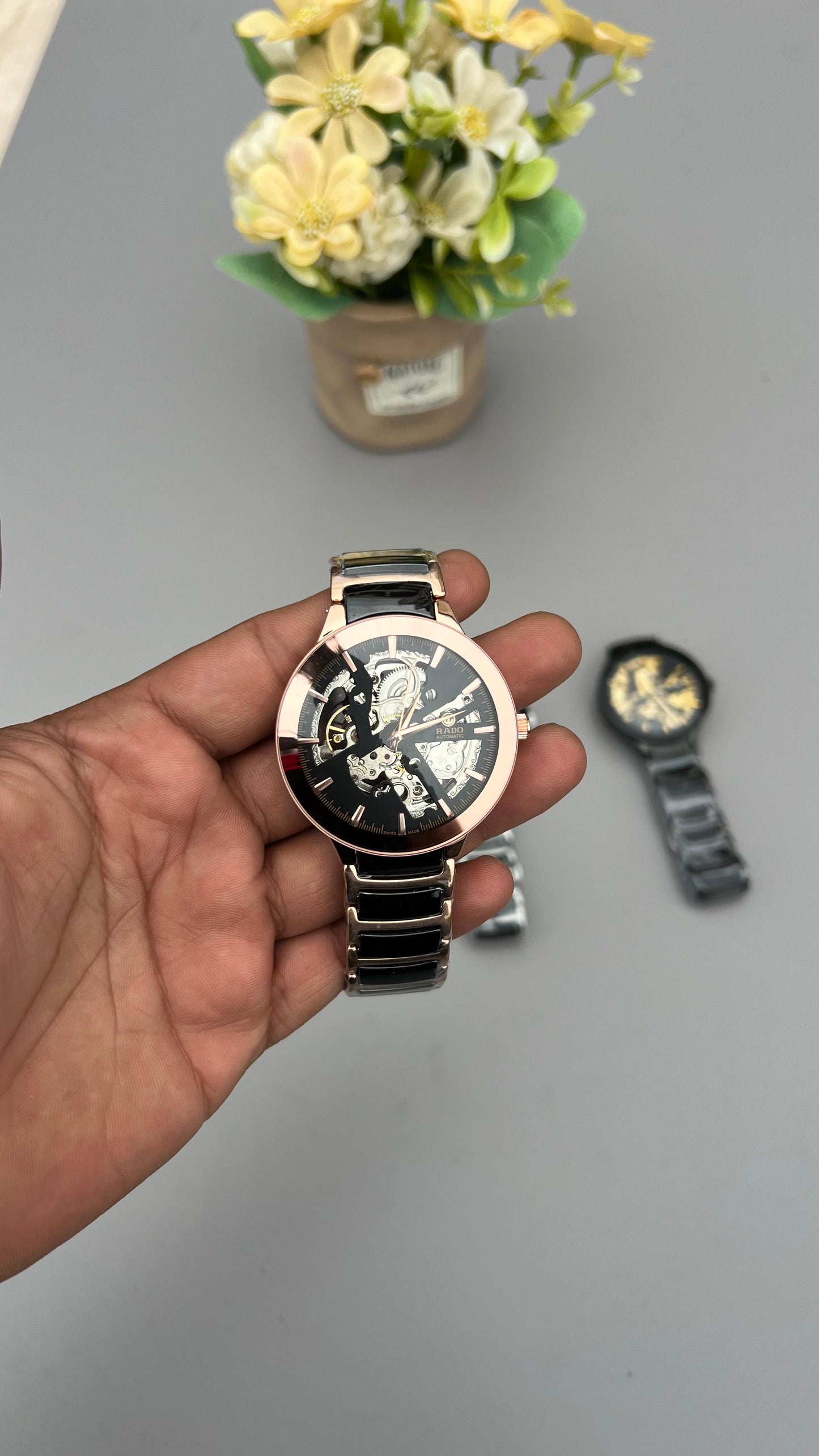 Hand holding a black and rose gold watch with a blurred background of flowers and another watch.