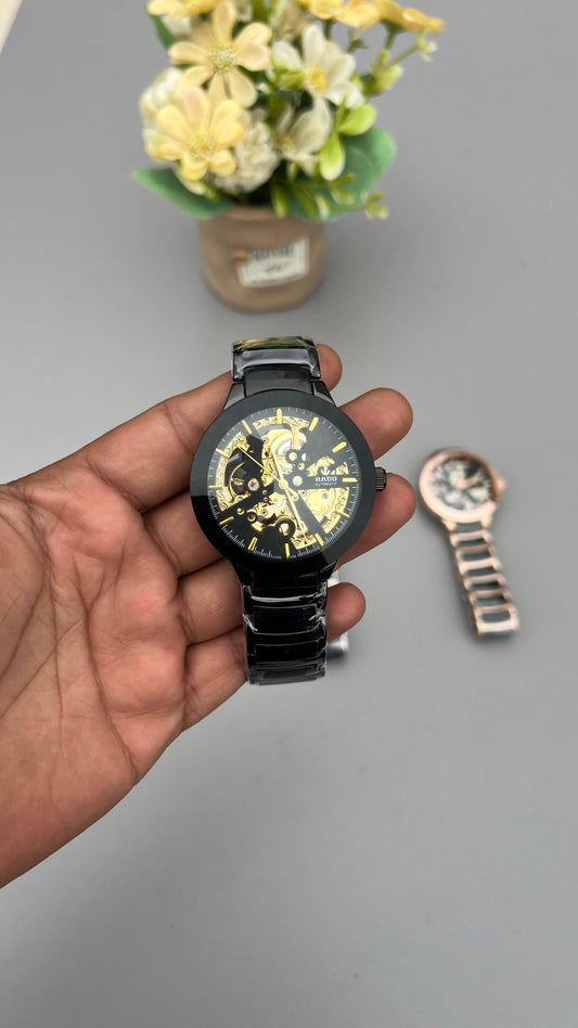Hand holding a black watch with yellow details on a gray surface with a small plant in the background.